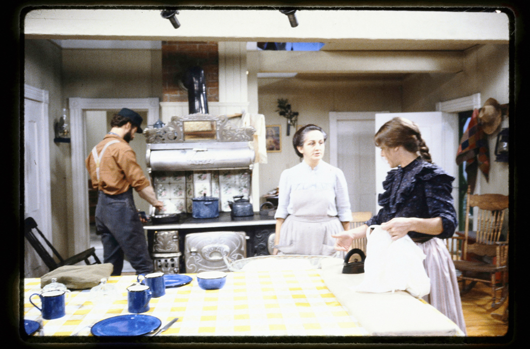 Dans la cuisine de Rose-Anna, Valérien Lavoie (Jacques L'Heureux) fait à manger sur la cuisinière au bois pendant que Rose-Anna St-Cyr (Nicole Leblanc) discute avec sa fille Juliette (Katerine Mousseau) qui repasse du linge.